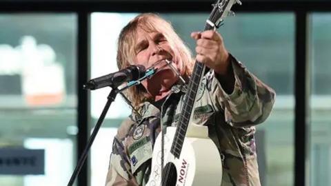 Mike Peters who has long blonde hair and is wearing a khaki camouflage jacket. He is stood at a microphone stand and is holding a cream guitar which he is playing and looking at. Behind him are some large windows and a man wearing a baseball cap.
© Nicholas Hunt/Getty Images