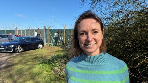 A woman in a blue jumper with green stripes. She is standing outside RAF Fairford and is smiling at the camera. There are cars and people behind her. It is a sunny day.