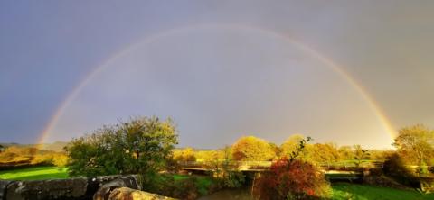 A rainbow semi-circle. The sky is filled with grey cloud. The right side is lit up by sunlight. There are trees in the foreground and background. 