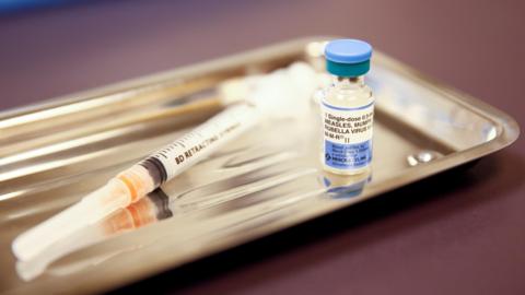 A stainless steel tray holding a glass vial labelled 'single does measles, mumps, rubella virus' and a hypodermic needle