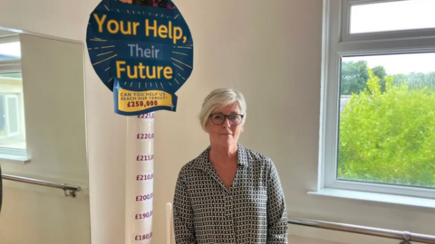 A woman standing next to a sign which looks like a lollipop - it says at the top 'Your Help, Their Future' and a £250,000 target. The pole can be seen from £180,000 and goes up in £10,000 increments until it reaches target. 