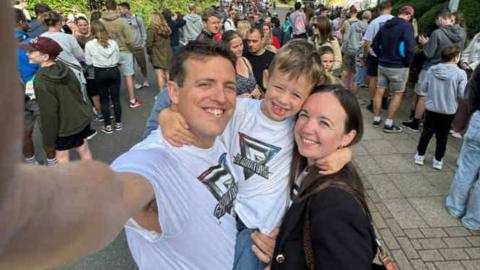 Jessica, a woman with dark hair smiles with her young son with light hair in the middle and her husband with dark hair.