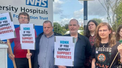A group of six people from Unite the Union outside Addenbrooke's Hospital in Cambridge. They are holding placards saying honk if you support support workers. A sign saying NHS can be glimpsed on the left behind them. 