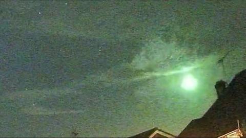A meteor bursts in a pale green fireball above a roof