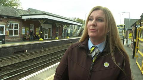 Station manager, Sonya Hampson wears a brown jacket with a yellow Merseyrail badge and a blue blouse and lime green, grey and light grey scarf. She has long blond hair. She is standing on the station platform.