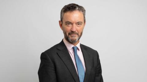Matthew Jowitt is smiling and looking at the camera in front of a grey background. He has short grey hair and a beard and is wearing a black shirt over a pink shirt with a blue tie. 