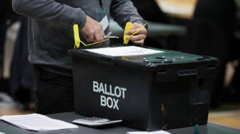 A black ballot box being opened by a man at a table. Just the man's lower body and hands are visible. He is using scissors to cut a yellow plastic tag which had secured the ballot box's lid.