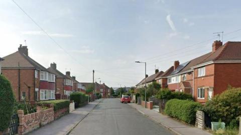 A suburban street lined either side with red brick local authority-type properties. Each has a small front garden abutting the empty road, most feature priver hedges.