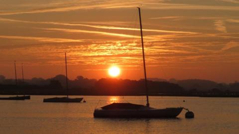 The Sun rises in an orange sky with boats on the river in front in silhouette