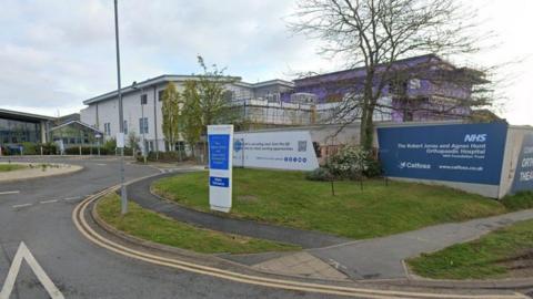 The entrance to an NHS hospital, which has blue signage with white writing indiciating it is The Robert Jones and Agnes Hunt Orthopaedic Hospital. There is a pavement bounded by grass verges, and road markings at a junction as wekk as double yellow lines.