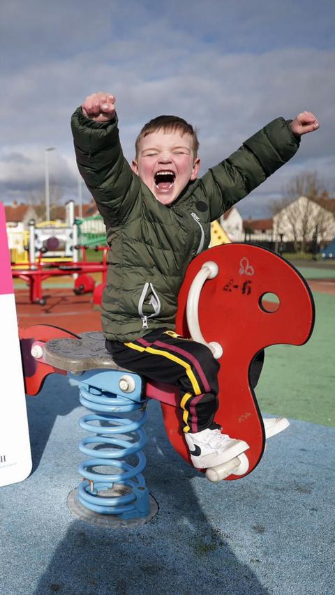 Dáithí in a green coat and black tracksuit bottoms. He is sat on a springy play park horse with his arms aloft. 