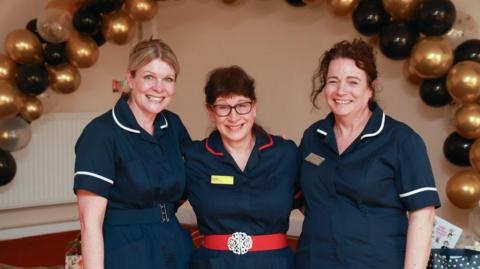 Catherine Draycott, Hazel Hawkins-Dady and Emily-Bloomfield: Three women in dark blue nurse uniforms are stood together with an arm around each other and smiling at the camera. In the background there are gold and black balloons in an arch.
