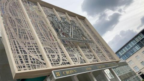 The front of Plymouth Crown Court. It is a modern building, featuring an intricate geometric pattern on the front made of metal and concrete, with colourful sections of glass or panels in the design. It also bears a coat of arms and a sign saying "CROWN AND COUNTY COURTS".