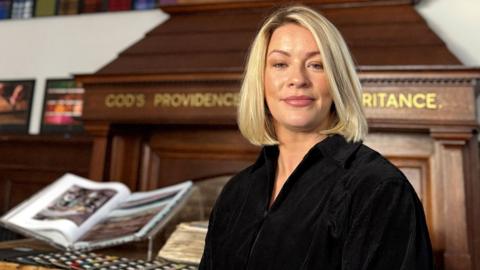 Fiona MacKenzie has blonde hair to her shoulders and is wearing a black top. She is sitting in a large room at Harris Tweed Authority offices. 