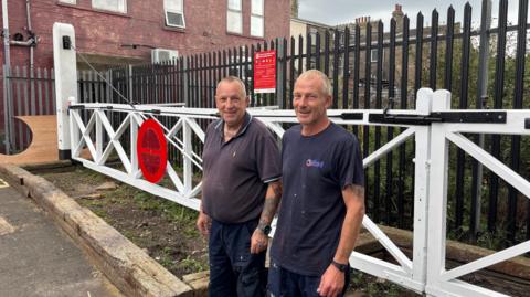 Two men wearing navy-coloured clothing and standing in front of the railway gates. They are both looking at the camera and smiling.