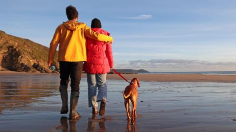 Couple walk their dog on a beach at sunset. They are both wearing colourful coats.