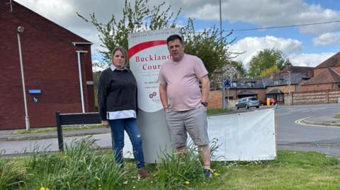 A man with brown hair and wearing a pale pink T-shirt and grey shorts is pictured standing next to his wife on a grass verge by the side of a road. The woman has blonde hair and is wearing a dark grey jumper and dark blue jeans. Both are standing in front of the 'Buckland Court' care home sign on the road side with houses and parked cars in the background.