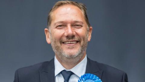 A man with short dark hair smiles. He is wearing a business suit and a blue rosette. 