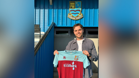 Love Island star Harry Cooksley pictured holding the Farnham Town Football Club after signing for the club. Harry is wearing a white t-shirt and grey shirt, and is holding a maroon and blue shirt. The Farnham Town FC crest can be seen in the background.