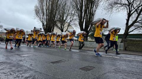 A group of people running on a road carrying bags on coal on their backs. They are wearing yellow t-shirts and black shorts. 
