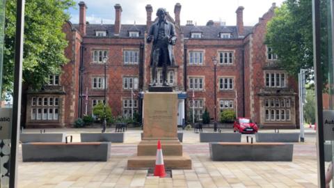 A bronze statue of a man in a long coat holding a pot. It stands on a stone plinth that has JOPSIAH WEDGEWOOD inscribed on it. There are benches either side of the statue and a large hotel behind.
