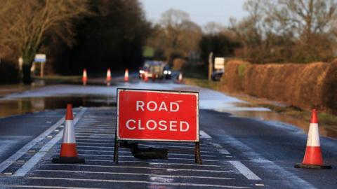 A road closure sign in a flooded road. The road also has cones on it.