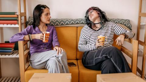 Two women holding mugs sit on a sofa with cardboard boxes in front of them, and shelves either side of them, with some books on the shelves.