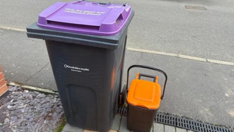 A black bin that says 'Lincolnshire recycles' on it in small white font with a purple lid that says 'Only dry paper and cardboard please'. Next to it is a small black caddy with a purple lid. It is situated at the end of a driveway next to a grey footpath and road.