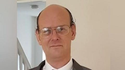 A man with a bald head and large, wire-framed glasses looks at the camera. There is a light reflected in the lenses of his flasses. He has brown, slightly curly hair at the sides of his head. He is wearing a white collared shirt and a dark jacket, which is not fully visible at the bottom of the photo.