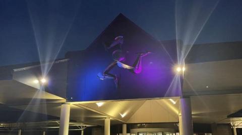 The exterior of the roof of the entrance to Noble’s Hospital, which features a triskelion sculpture that has been is lit up with blue and pink lights.