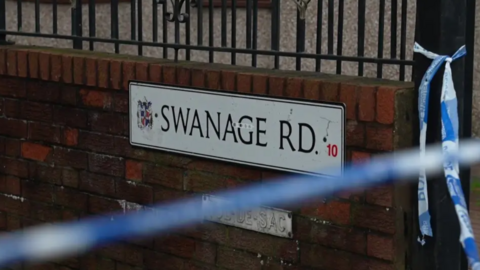 A close up of road sign on a brick wall, which says Swanage Road. There is police tape near the sign