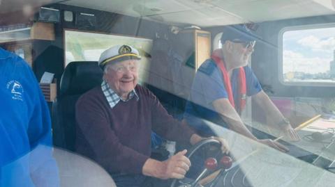 Roger Barnham, wearing a sailor's hat, is sat holding the wheel of a vessel.
