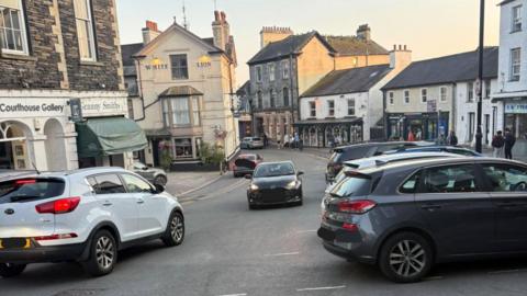 A black car being driven the wrong way up the main street in Ambleside, Lake District. There are cars driving in the oncoming direction. There are lots of shops and buildings which include pubs, outdoor shops and a gallery.