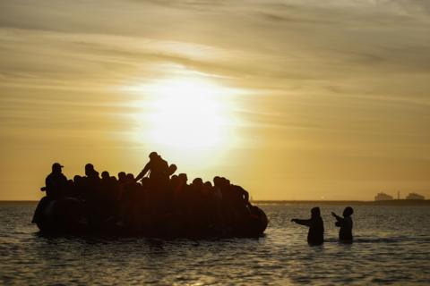 A silhouette of a dinghy carrying multiple people with two would be passengers standing in the water trying to board
