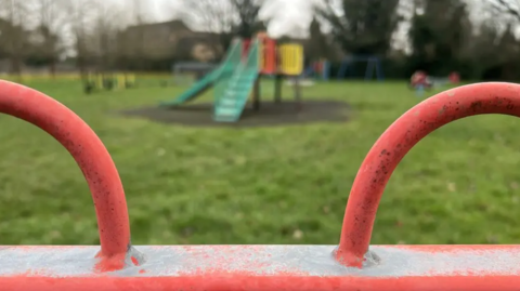 A children's play area with a green slide in the background and a red metal fence in the foreground.