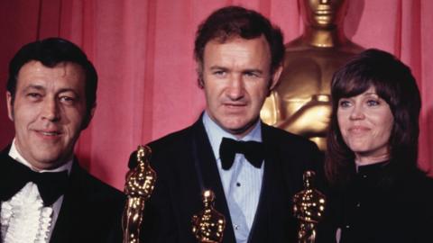 Gene Hackman, wearing a tuxedo, holds his Oscar alongside actress Jane Fona and Philip D' Antoni, a producer. The photo is from 1972. A giant Oscars statue stands behind them