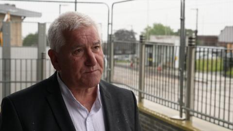 A man wearing a white shirt and black blazer with short grey hair looks at the camera. Behind him are grey metal fences on a train platform. 
