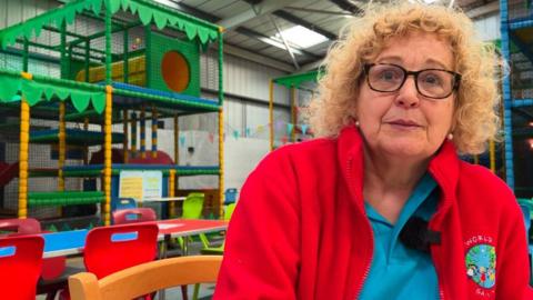 Jane Coulson is wearing a red jacket with a blue shirt underneath. She has curly blonde hair and is wearing black glasses. She is sat in front of play equipment, tables and chairs.