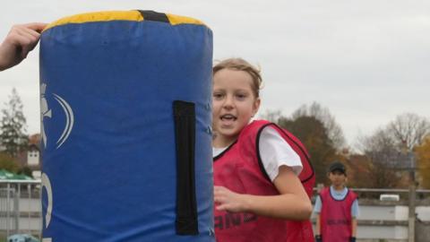 A primary school-aged girl in a pink tabard over a white T-Shirt running towards and about to make contact with a blue tackle bag, an upright foam pad about 1.5 metres (4.9ft) tall. Behind her is another, similarly-dressed pupil. They are on the grounds of a rugby pitch.