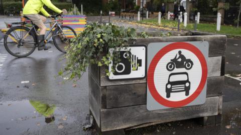 LTN box with a plant, camera sign and a red symbol with car and motorbike inside in the foreground. Behind, a cyclist goes past