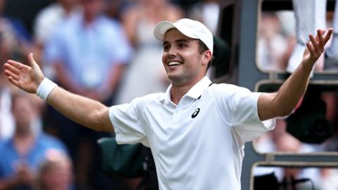 Arthur Fery, 23, in tennis whites celebrates with both hands in the air with his palms facing upwards.