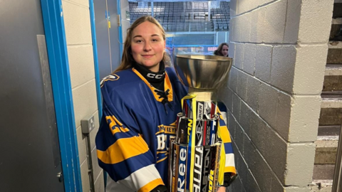 A woman in a blue, yellow and white ice hockey jersey. She is holding a large trophy inside a stadium.