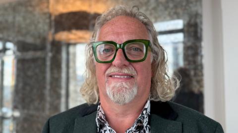 Michael Stewart stands, looking at the camera. He has long curly hair, and is wearing a shirt and a green suit jacket. Behind him is an ornate-looking marble wall. Wearing green glasses. 