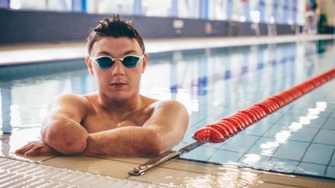 Lyndon Longhorne, with short black hair and swimming goggles, is propping himself on the edge of the swimming pool.