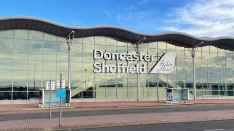 DSA terminal. A large glass fronted building with a wavy roof. With Doncaster Sheffield written in large white letters alongside a smaller sign saying Robin Hood
