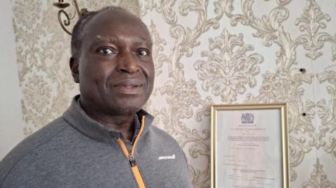 Nelson Shardey has short black and grey hair and is wearing a grey top with an orange zip. He is stood in his living room next to a framed certificate of naturalisation issued by the Home Office.
