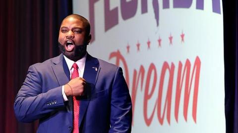 Bryon Donalds holds his hand to his chest as he takes to a stage following his victory in a primary election contest