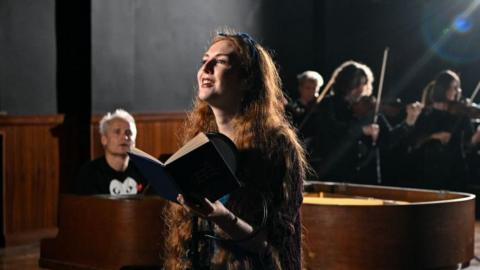 Len Pennie, a woman with long red hair, speaking into a microphone in a recording studio while holding a book in front of her. String players are visible in the background. Paul Leonard-Morgan is sitting at a grand piano behind her. He has blond hair and is wearing a T-shirt.