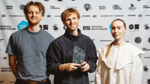 Three members of redolent, two male and one female, stand holding their award trophy in front of a wall of sponsor logos