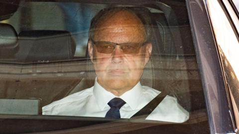 A close-up of man sitting in the passenger seat of a car. We can see his face and shoulders. He is wearing sunglasses and a white shirt with a black or navy blue tie.